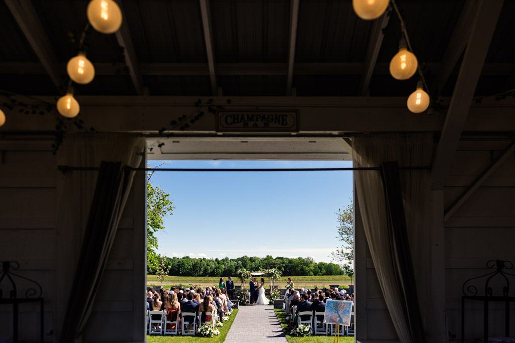 View from inside a barn at Être Farms looking out at an outdoor wedding ceremony on a sunny day, with guests seated in rows