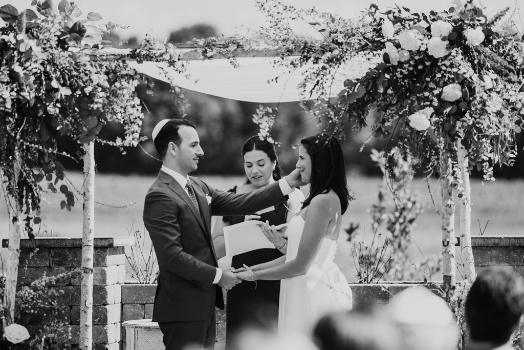Black and white photo of groom brushing a hair from the bride’s face during an outdoor wedding ceremony under a chuppah at Être Farms