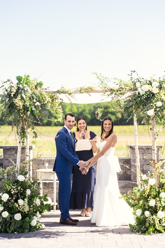 A bride and groom hold hands and smile during an outdoor wedding ceremony with floral decorations at Être Farms