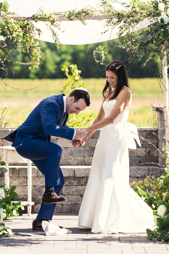 Groom in a blue suit about to break glass underfoot as the bride smiles during an outdoor wedding ceremony at Être Farms