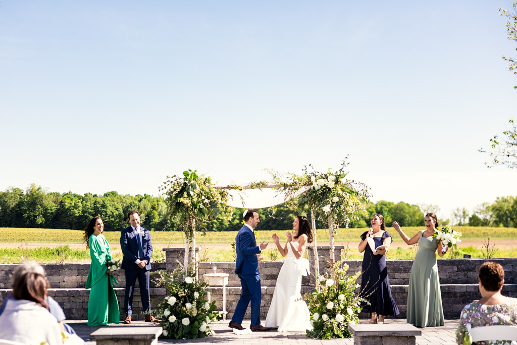 Newlyweds celebrate after breaking the glass during their Être Farms outdoor wedding ceremony with friends on a sunny day