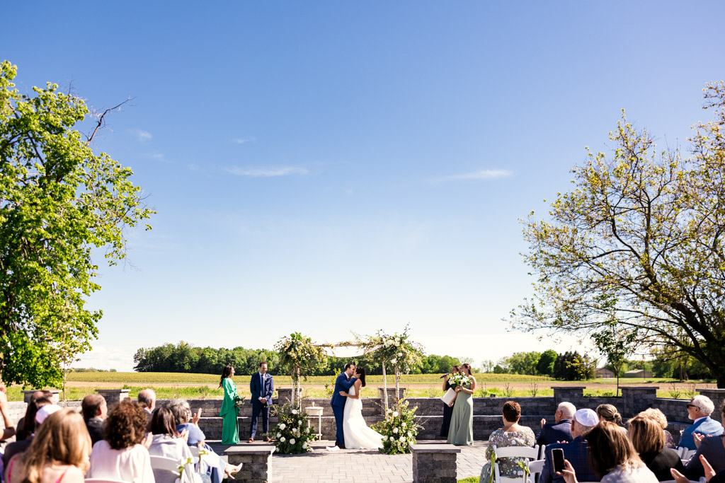 Newlywed couple kisses at an outdoor Être Farms wedding ceremony under a clear blue sky, surrounded by guests and lush greenery