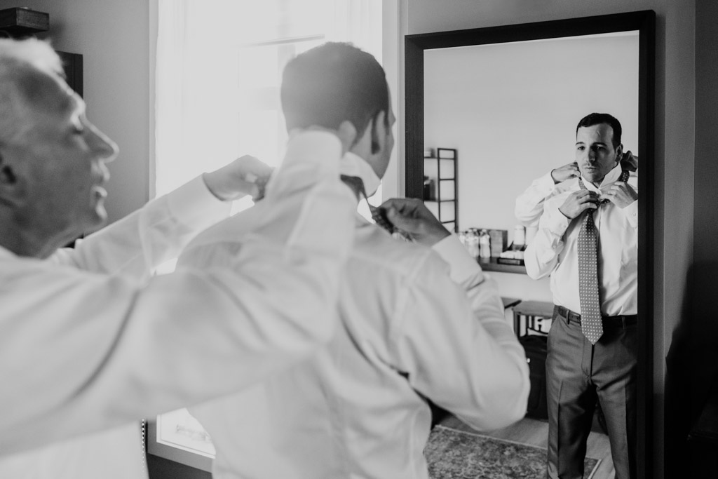 Black and white photo of groom's father man helping groom adjust his collar as he ties his tie in front of a mirror while getting ready at Être Farms