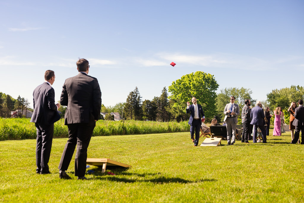 Guests in suits playing cornhole on a the Être Farms lawn, with others socializing in the background during spring wedding reception