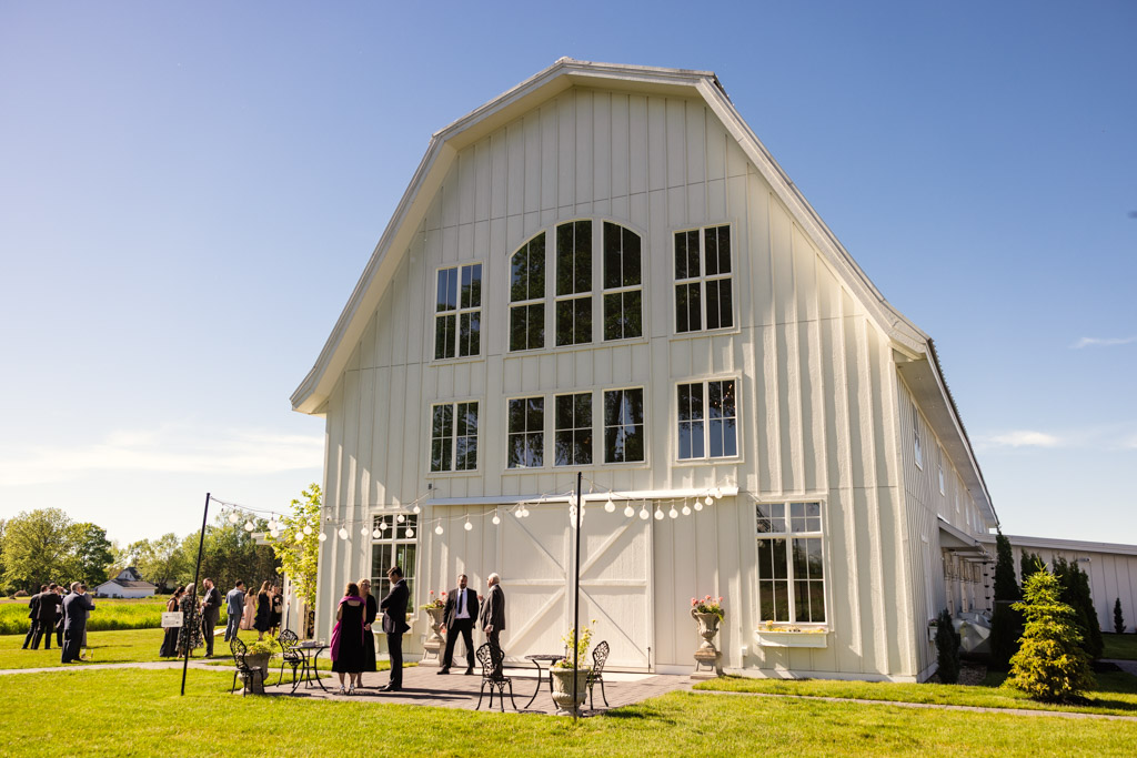 Wedding guests outside the large white Être Farms barn venue on a sunny day, with string lights and patio seating