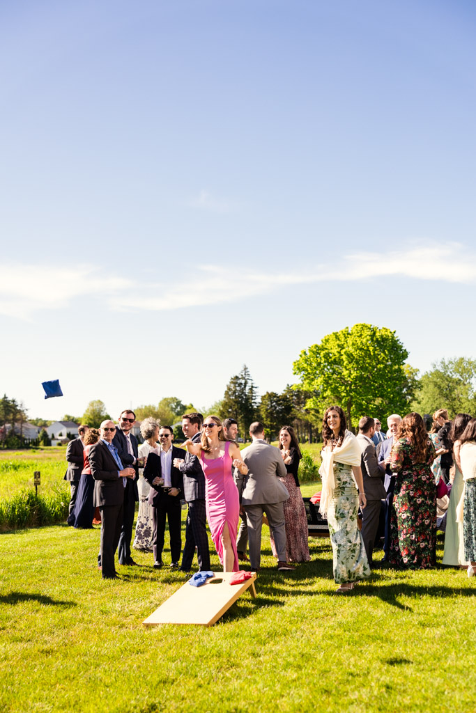 Guests in formal attire play a game of cornhole on the sunny lawn at Être Farms during spring wedding reception