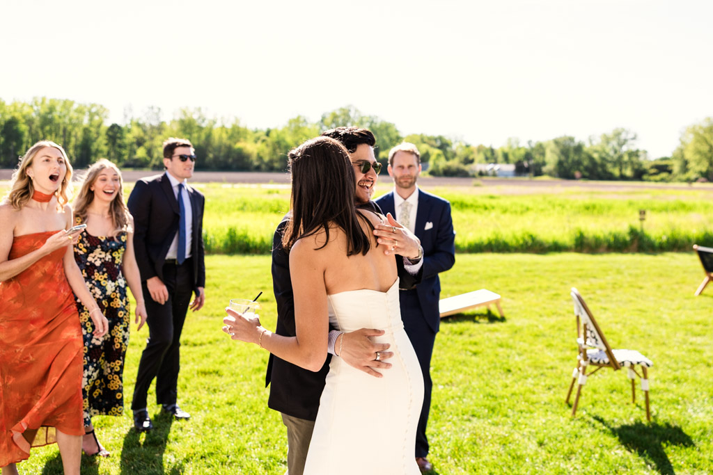 Bride hugs a guest outdoors at Être Farms while others in colorful dresses and suits smile during cocktail hour at Être Farms wedding reception