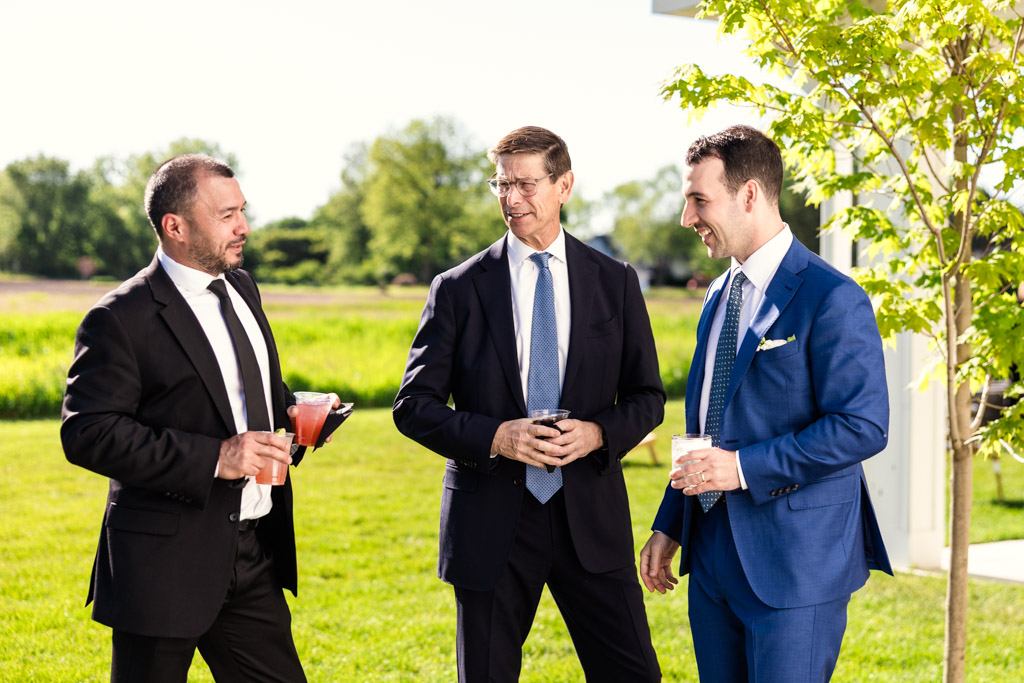 Groom speaks with two guests as they stand outside on the grass at Être Farms, holding drinks on a sunny day