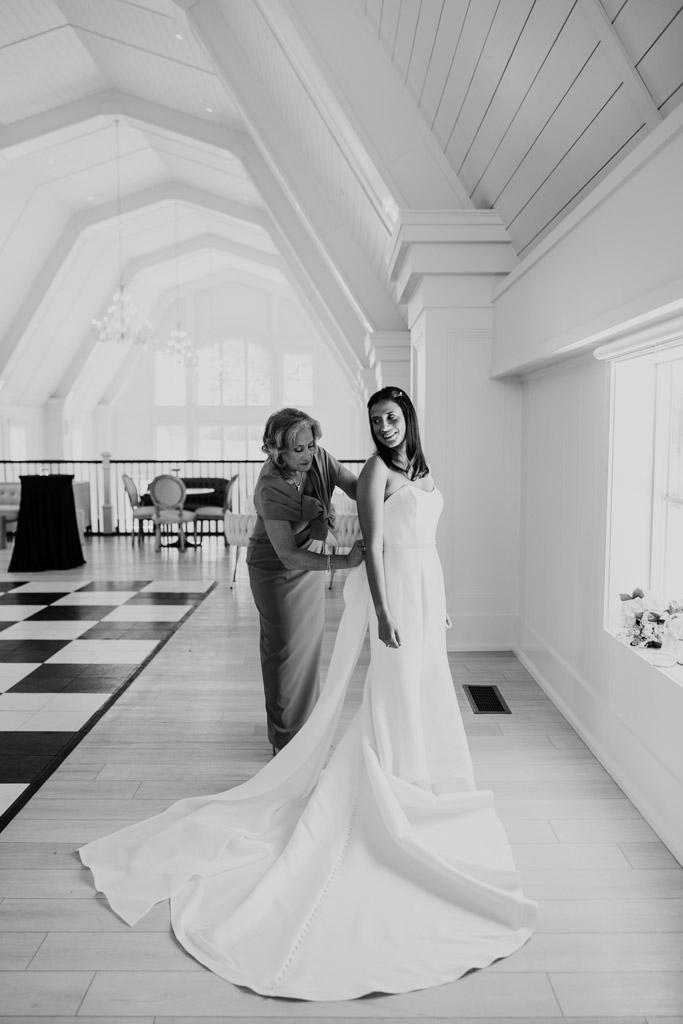 Black and white photo of bride's mother helping bride adjust her dress in a bright room with high ceilings at Être Farms