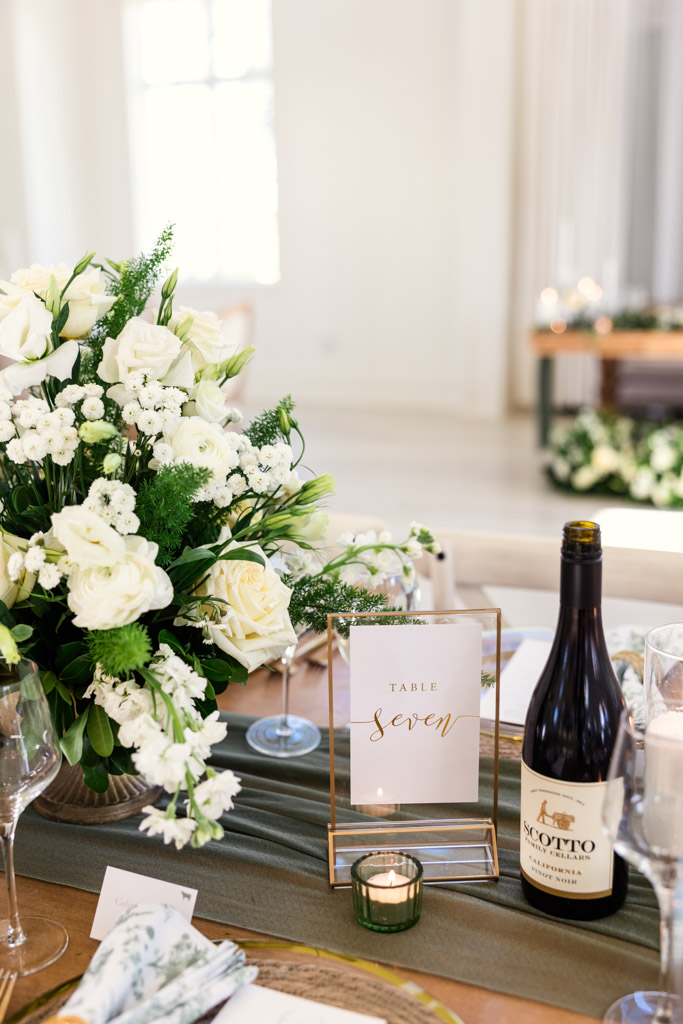 Guest table setting with white flowers, a wine bottle, and a table sign in gold script for Être Farms wedding reception