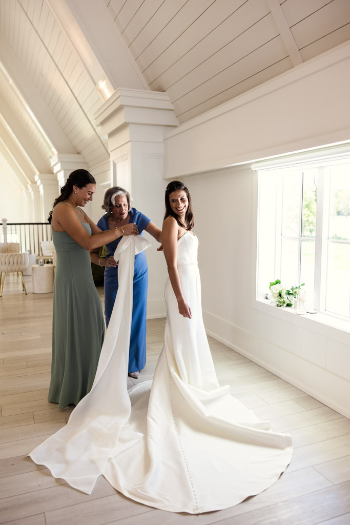 Bride in white dress smiles as her mother and sister help arrange her train by a window in a bright room at Être Farms