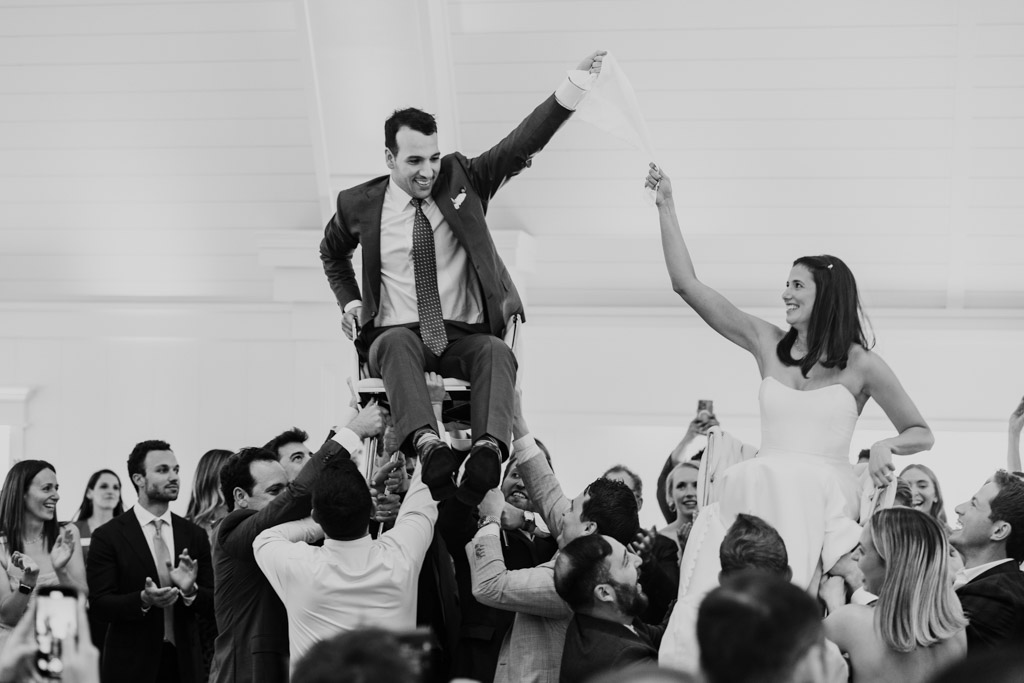Black and white photo of bride and groom lifted on chairs by guests during the hora at their Être Farms wedding celebration