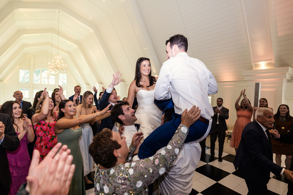 Bride and groom are, lifted on chairs by guests during the hora, surrounded by cheering friends at Être Farms wedding reception