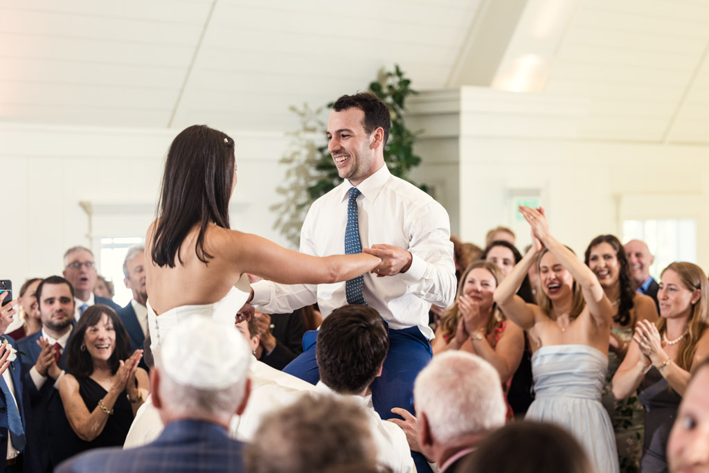 Happy bride and groom hold hands while they are lifted on chairs during the hora at their Être Farms wedding reception