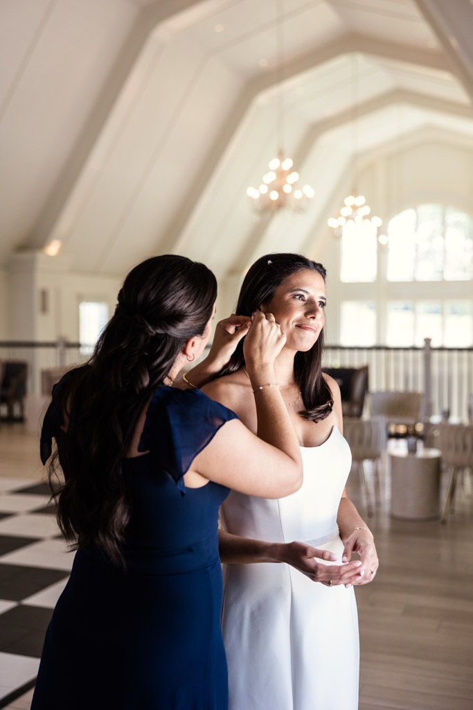Bride's sister helps bride adjust her earring in a bright, elegant room with large windows and chandeliers at Être Farms