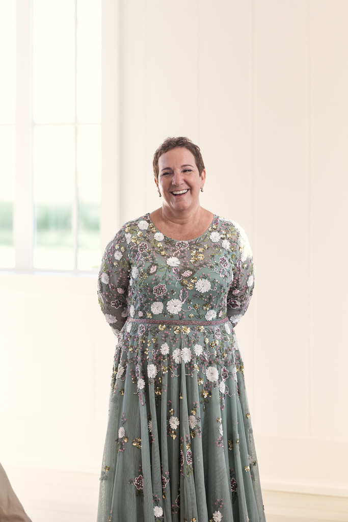 Mother of the groom in a long, floral dress stands smiling in the bright Être Farms wedding reception space with large windows in the background