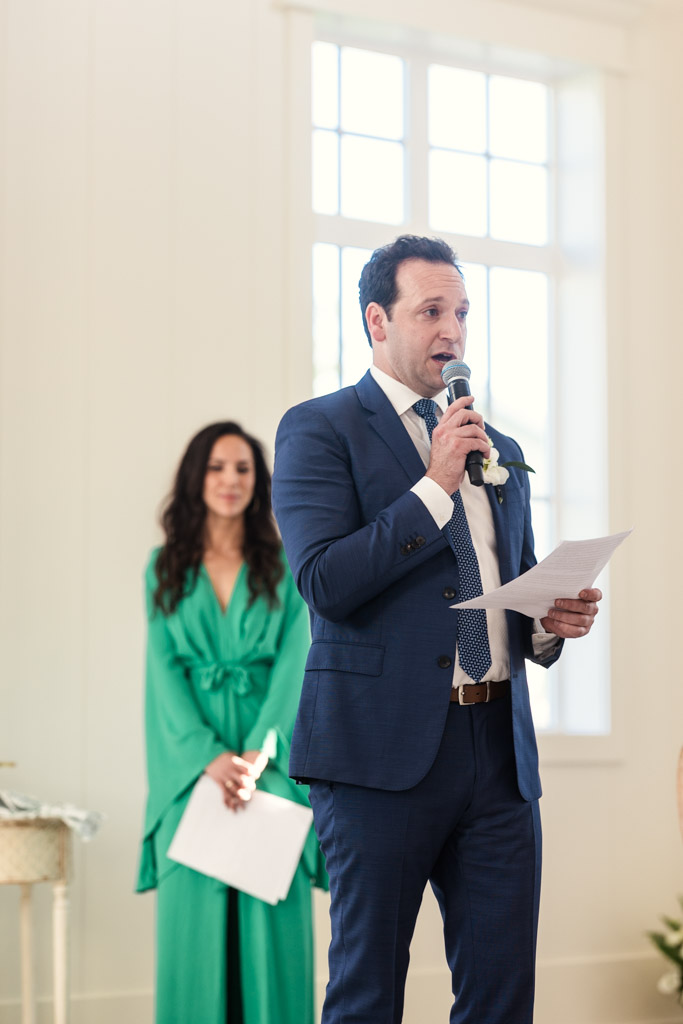 Best Man in a blue suit gives speech during Être Farmswedding reception while groom's sister dressed in green stands behind him