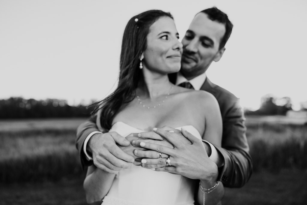 Black and white photo of couple embracing outdoors at Être Farms, smiling at each other, with focus on their wedding rings
