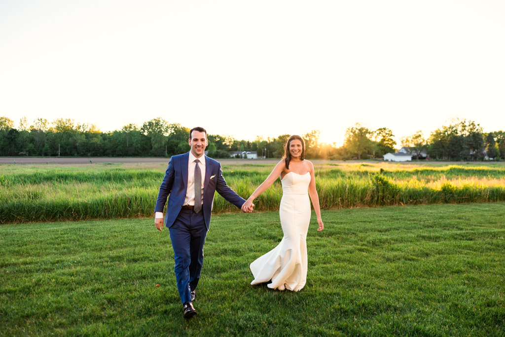 Bride and groom holding hands and walking on grass at sunset with lush greenery in the background at Être Farms