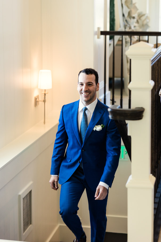 Groom in a blue suit smiling while walking up a staircase during the first look of his Être Farms wedding celebration