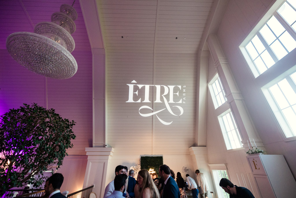 People mingling in a bright, elegant room with a chandelier and a large Être Farms logo projected on the white wall inside the venue's reception space
