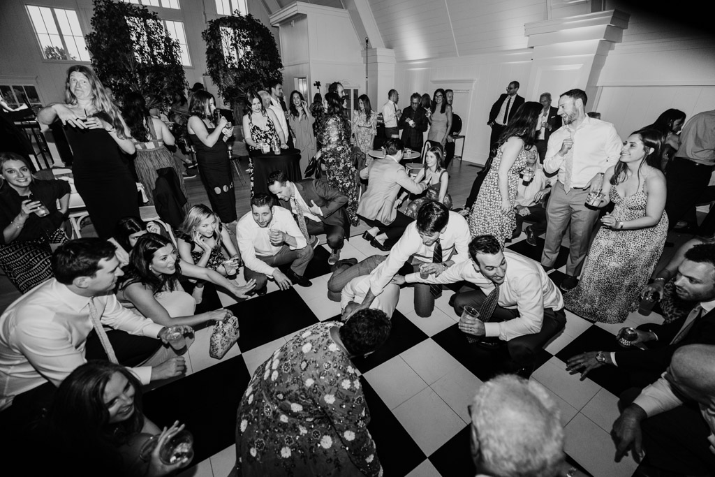 Black and white photo of groom and guests dressed up, dancing and crouching on a black and white checkered floor at an Être Farms wedding reception