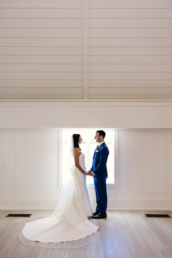 Bride and groom stand holding hands, facing each other in front of a bright window in a white room at Être Farms