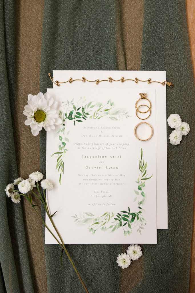 Wedding invitation with floral design, gold rings, a bracelet, and white flowers on green fabric for spring Être Farms wedding celebration