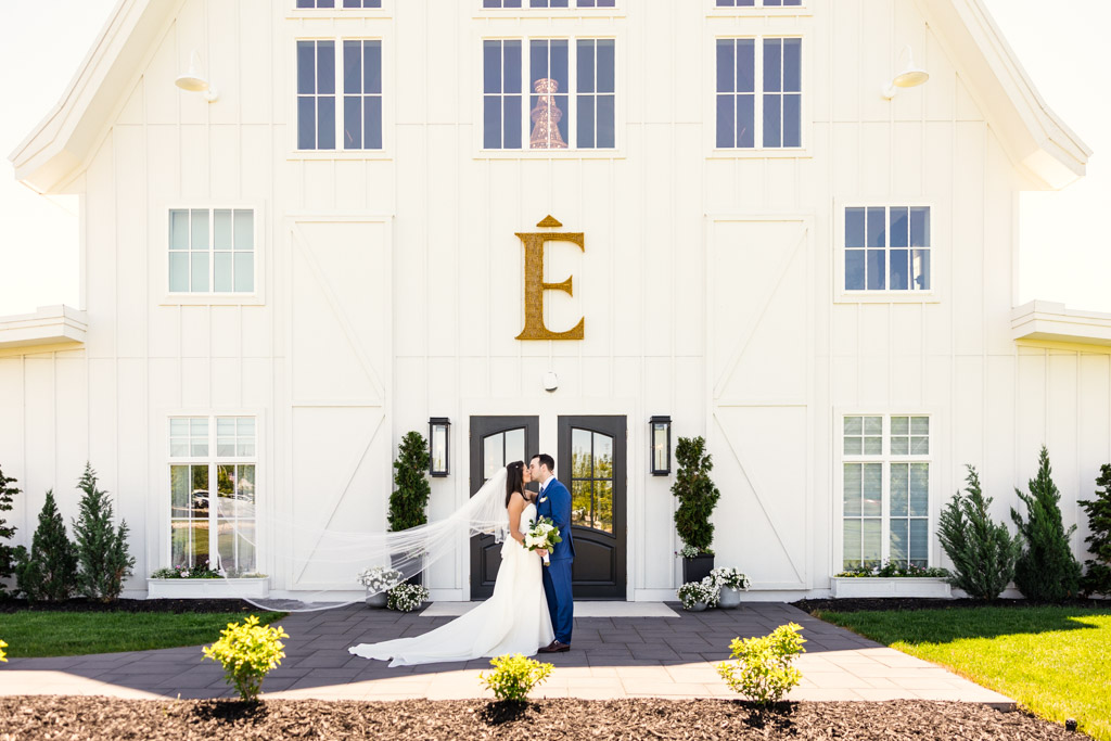 Bride and groom kiss outside the large white barn at Être Farms, with a gold letter