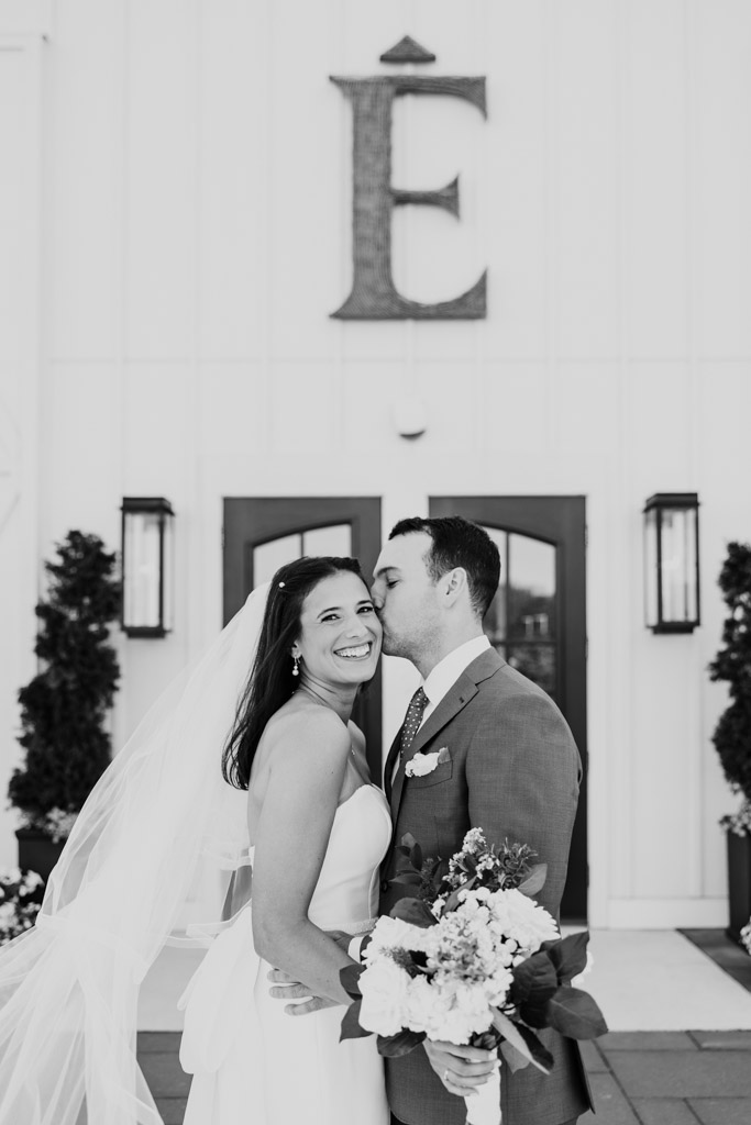 Black and white photo of bride smiling as groom kisses her cheek outside Être Farms main entrance