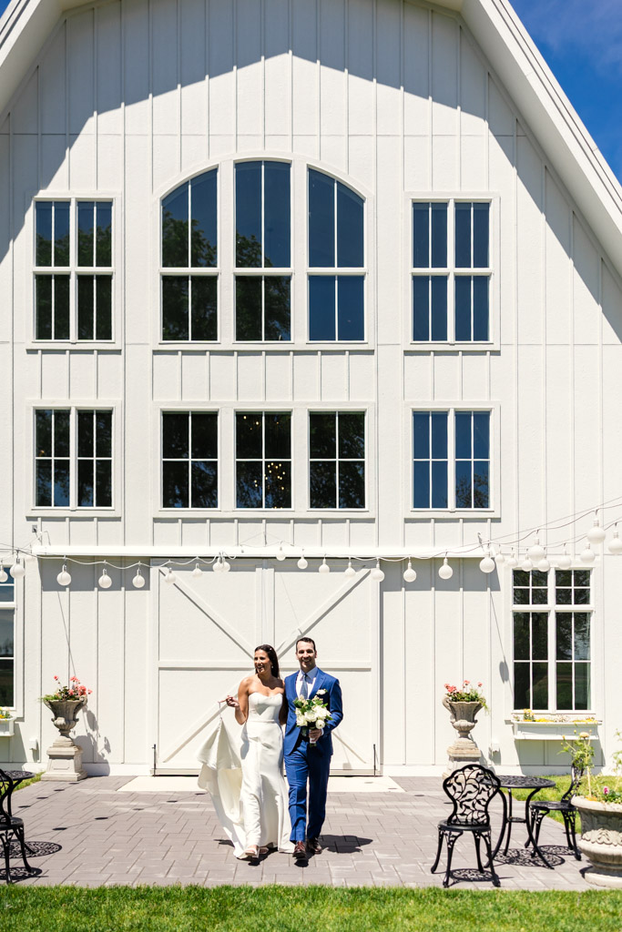 A bride and groom walk outside Être Farms’ white barn venue on a sunny day, holding hands and smiling