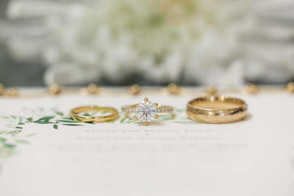 Detail photo of three gold rings, including a diamond engagement ring, displayed on a floral wedding invitation background for Être Farms wedding celebration