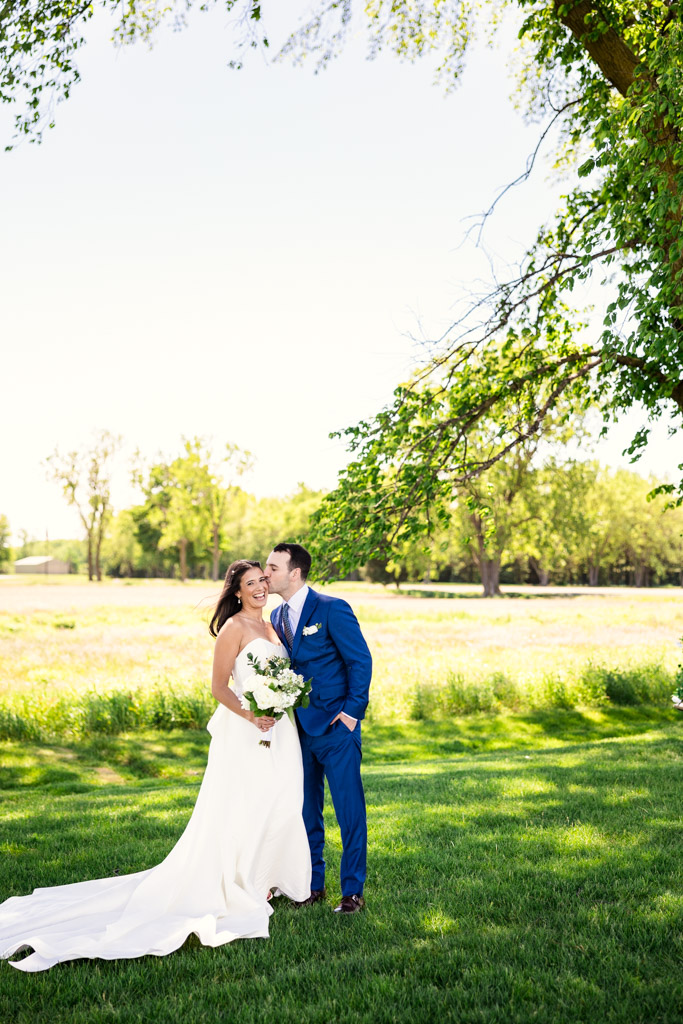 Groom kisses bride's cheek as they stand smiling together on the grass under trees at Être Farms, with a sunny field in the background