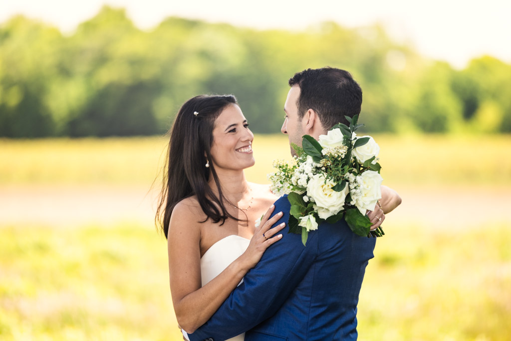 Bride and groom smiling at each other outdoors at Être Farms, bride holding her bouquet, a green field in background