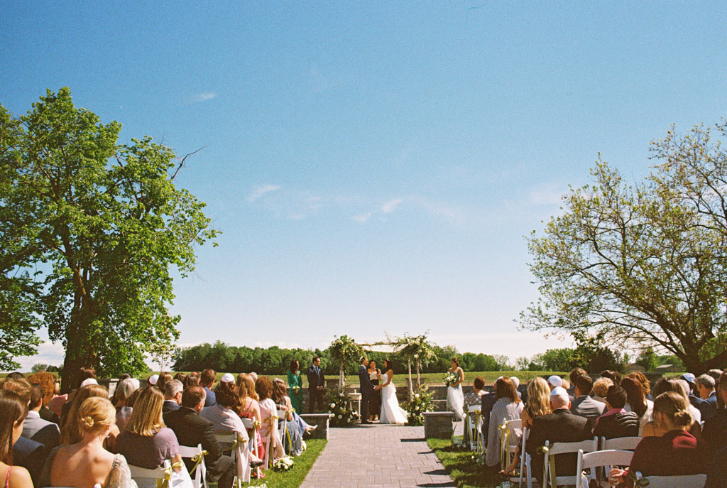 Film photo of outdoor wedding ceremony at Être Farms with seated guests, trees flanking each side, and a couple under a custom chuppah beneath a clear blue sky