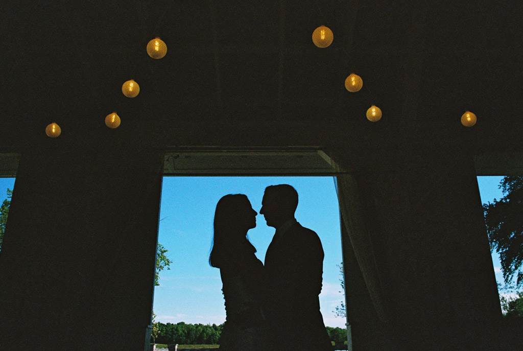 Film photo silhouette of a couple facing each other under hanging lights at Être Farms, with a bright blue sky in the background