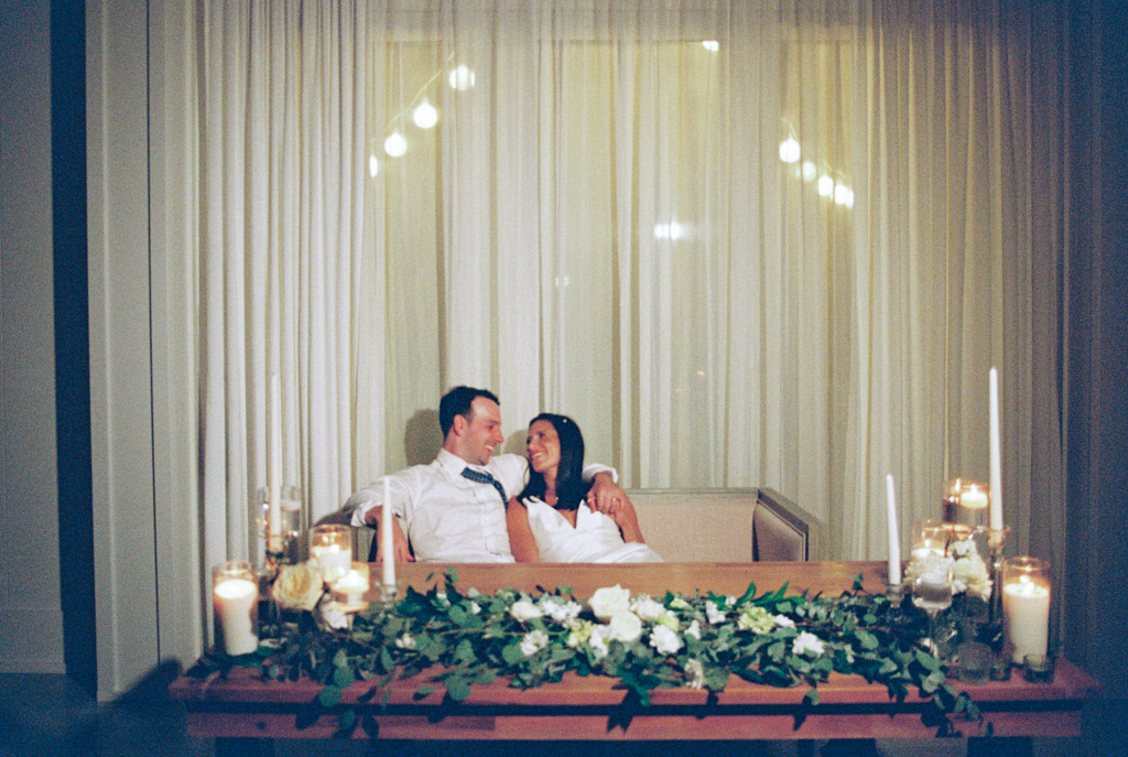Film photo of happy newlyweds at night, sitting together behind their head table decorated with candles in front of sheer curtains