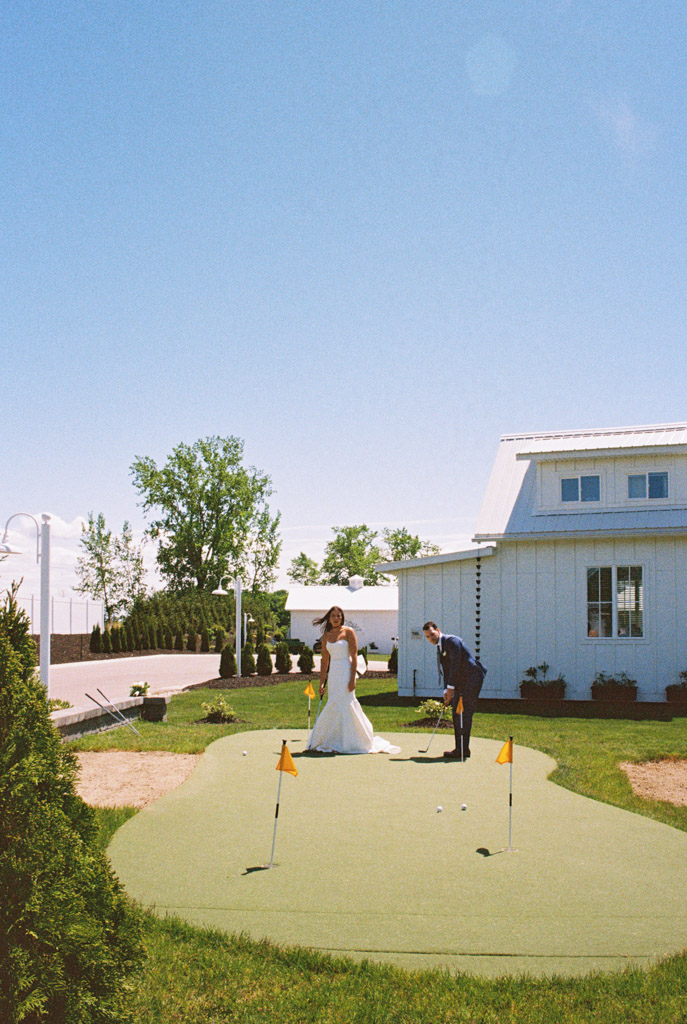 Film photo of bride and groom playing golf on a green lawn outside Être Farms under a clear blue sky