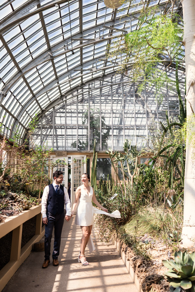 A couple in formal attire walks hand-in-hand inside the Desert Room during their engagement session at Garfield Park Conservatory, the woman twirling her dress