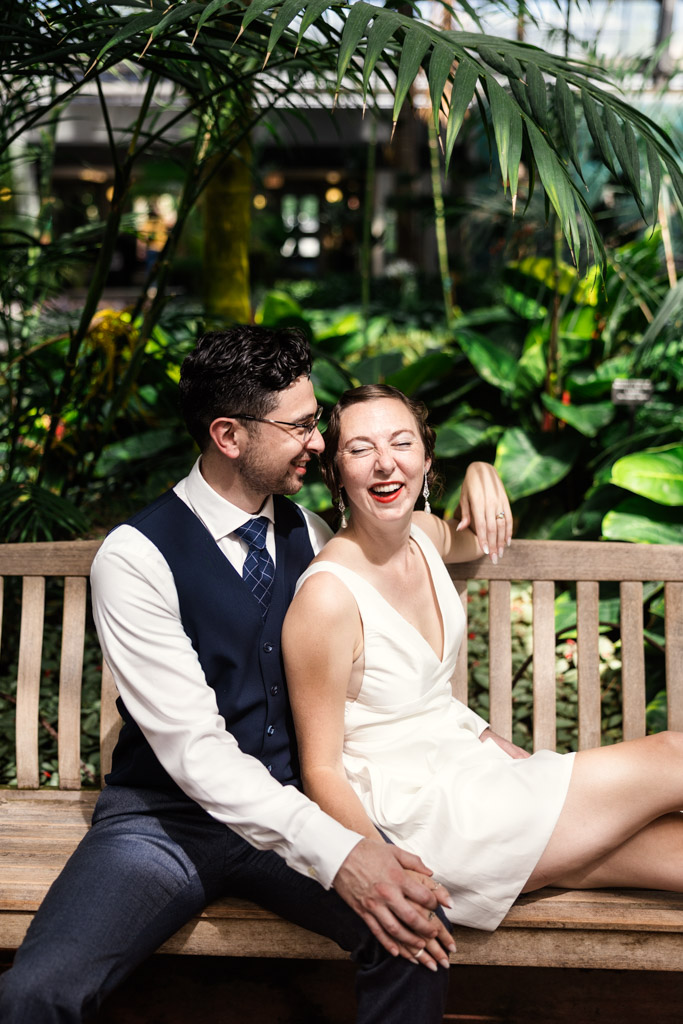Happy couple in formal 1920s-themed attire sits on a wooden bench, surrounded by lush green plants, during their engagement session at Garfield Park Conservatory