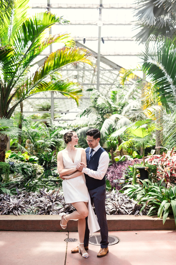 Man and woman pose together, smiling, in the bright, lush Garfield Park Conservatory filled with tropical plants during their 1920s-themed engagement session