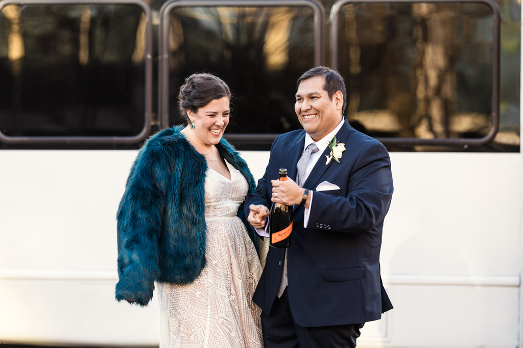 A smiling couple dressed formally stands outside, holding a champagne bottle together.