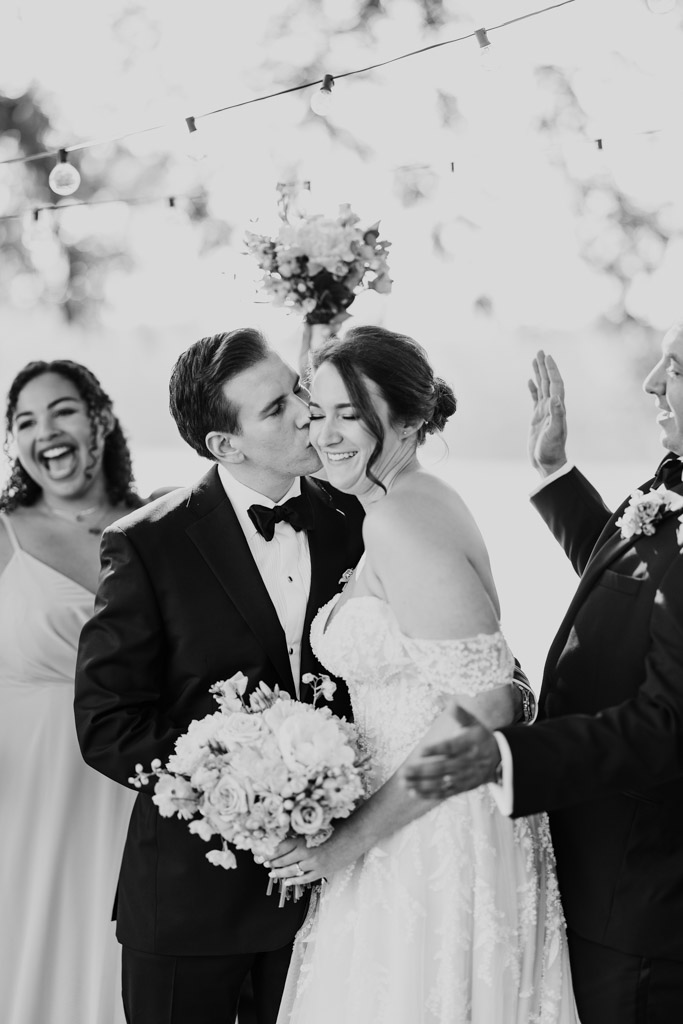 Black and white photo of groom kissing smiling bride's cheek at their Kemper Lakes Golf Club wedding ceremony with joyful guests around them