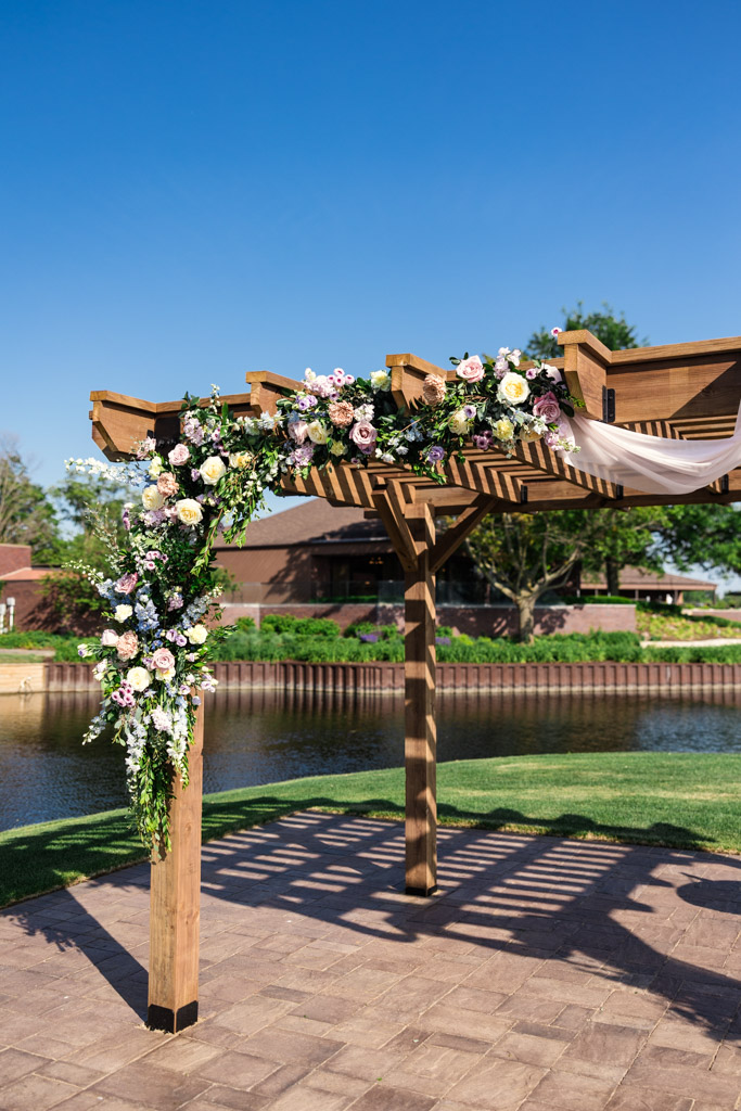 Wooden pergola decorated with flowers and fabric stands by a pond under a clear blue sky for wedding at Kemper Lakes Golf Club