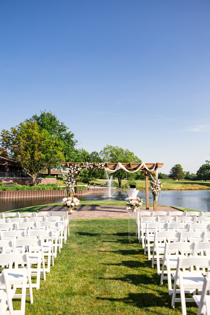Outdoor wedding ceremony space at Kemper Lakes Golf Club with white chairs, a floral arch, and pond view under a clear blue sky