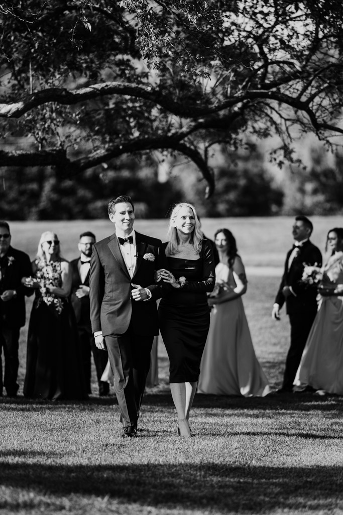 Black adn white photo of groom walking arm-in-arm with his mother at outdoor wedding ceremony at Kemper Lakes Golf Club