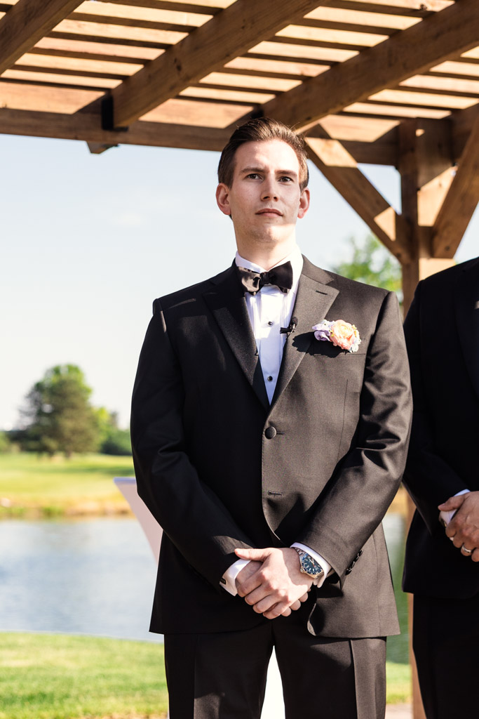 Groom in a black tuxedo standing outdoors under a pergola at Kemper Lakes Golf Club, with a pond and trees in the background