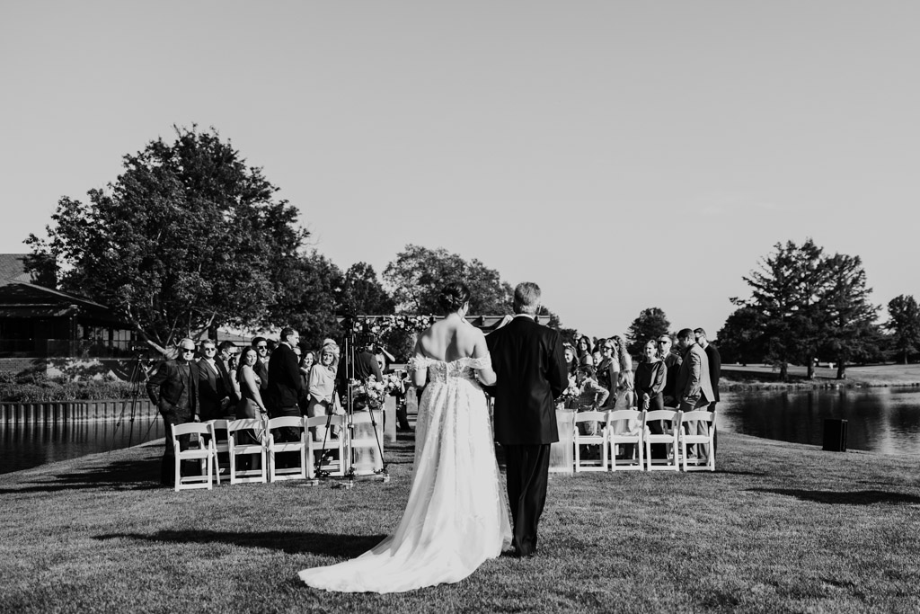 Black and white photo of bride and her father walking down the aisle during her Kemper Lakes Golf Club lakeside wedding ceremony