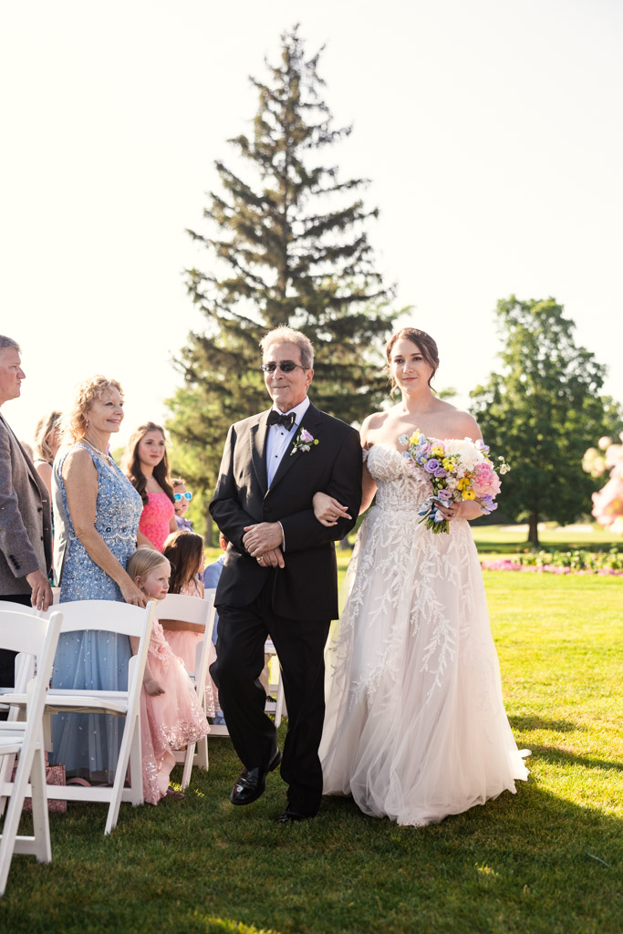 Bride in a white gown walks down the aisle with her father at Kemper Lakes Golf Club, surrounded by seated guests outdoors