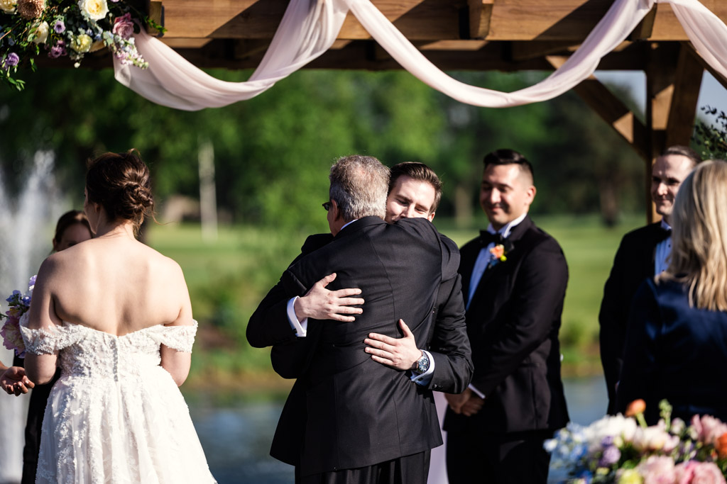 Groom hugs bride's father during his outdoor Kemper Lakes Golf Club wedding ceremony while guests look on and smile